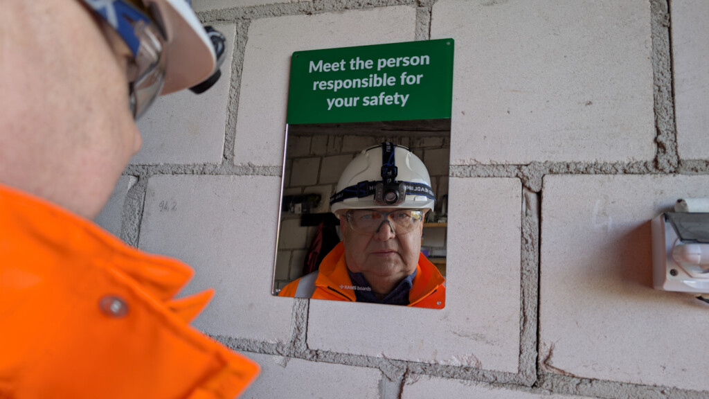 Un lavoratore edile con DPI guarda il proprio riflesso in uno specchio con la scritta "Meet the person responsible for your safety" montato su un muro di mattoni.