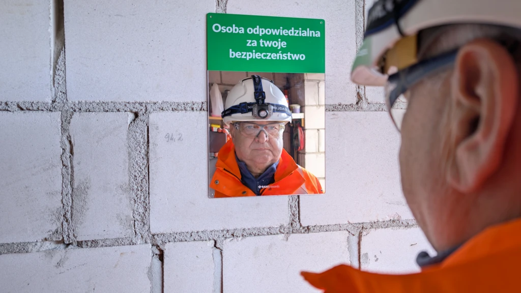 Lustro z napisem „Osoba odpowiedzialna za twoje bezpieczeństwo” zamontowane na ścianie z betonu komórkowego. W lustrze odbija się pracownik budowlany w odzieży ochronnej, patrzący na swoje odbicie.