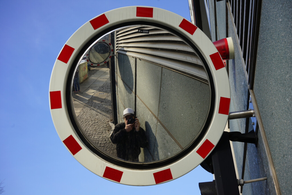 A round convex safety mirror with red reflectors mounted on a building, reflecting a photographer and the surrounding urban environment.