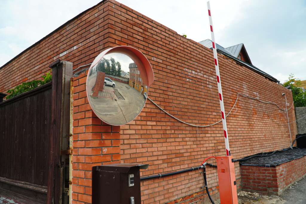 A convex traffic mirror mounted on a brick wall reflects a courtyard with parked cars and buildings, improving visibility around blind spots.