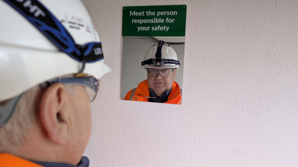 A construction worker in high-visibility orange gear and a safety helmet looks at a mirror with a sign above it reading, “Meet the person responsible for your safety.”